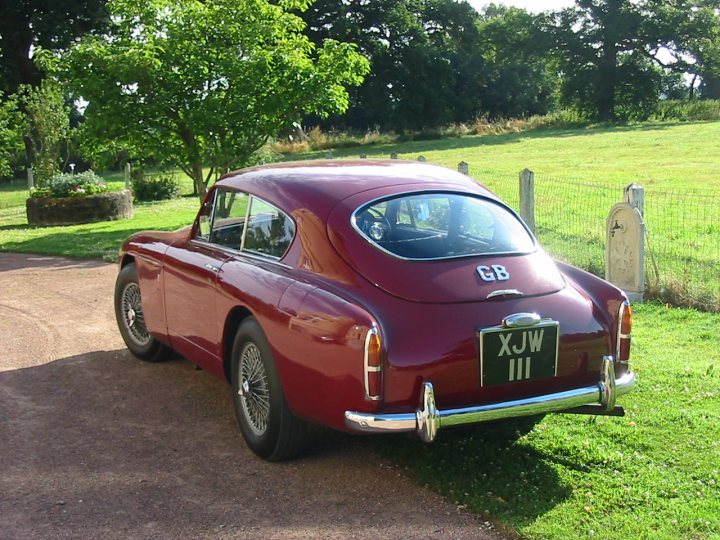 Stickers Paint Pistonheads Damage - This is an image of a classic car, specifically a 1957 Jaguar XJW 3.4, parked on a dirt path in a more rural setting. The car has a vibrant red paint job and features the iconic round rear side windows. In the background, there's a beautiful garden with a neatly maintained lawn and a tree. Around the car, there's a fence with a wooden gate indicating a private property or a gated driveway.