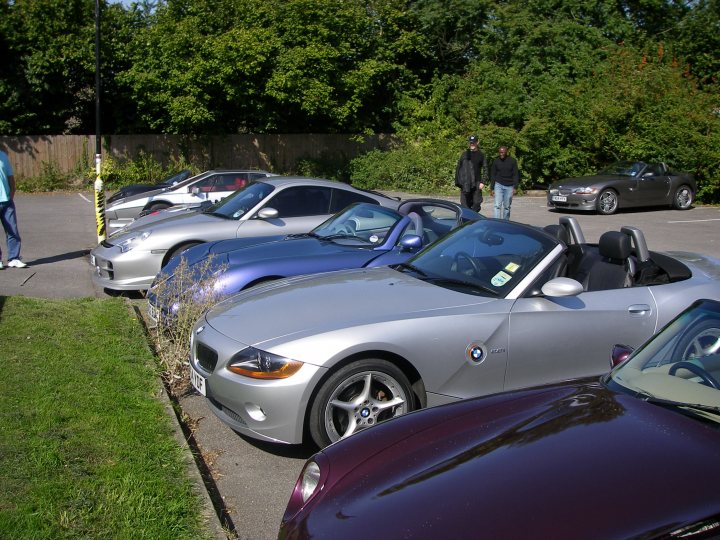 Pistonheads - The image shows a row of luxury cars parked next to each other in a parking lot. A man is standing near the front of the BMW, which is gray. There is a paved sidewalk alongside the cars, and grass is visible in some areas. Finally, there are trees in the background, suggesting a suburban or park setting.