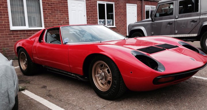 Miura SV being restored - Page 1 - Lamborghini Classics - PistonHeads - The image features a vintage sports car parked on a city street. The car, a vibrant red with a sleek design, has yellow accents and a distinctive black roof. It's parked in front of a brick building with white trim, which includes a front door and another, smaller door on its side. An SUV is partially visible in the background, contrasting with the vintage style of the sports car. The overall scene suggests a quiet urban setting.