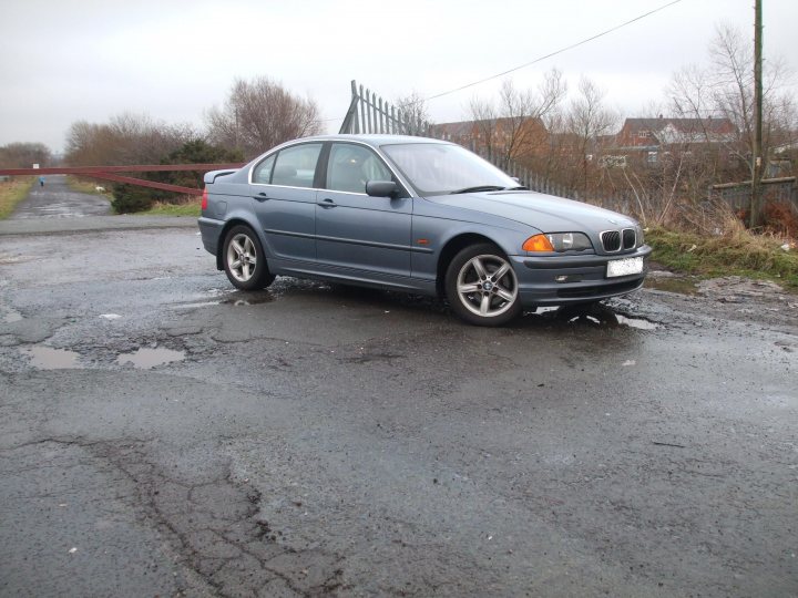 Pistonheads - The image features a gray BMW car parked on the side of what appears to be a wet, pothole-strewn road or parking lot. The car has black rims and a distinctive kidney design grille. In the background, there is a covered pavilion and a metal fence. The sky is cloudy, suggesting an overcast day. There are trees and shrubs in the vicinity of the car, indicating a semi-urban or suburban setting.