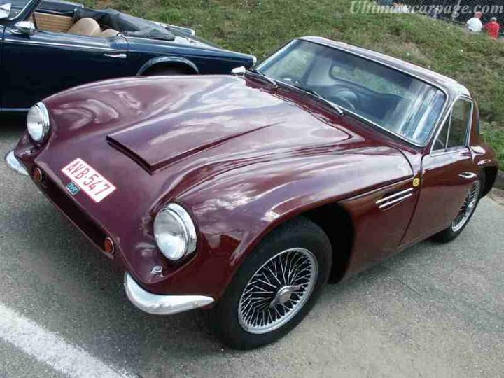 Early TVR Pictures - Page 35 - Classics - PistonHeads - The image presents a scene from across the street of an old-fashioned, maroon-colored car parked in a parking spot. The car's shiny finish and distinctive features, such as a prominent grill and round headlights, suggest it's an old Aston Martin. A license plate is visible at the front of the vehicle, and there's another car parked to its side, indicating the car might be part of a larger event or gathering.