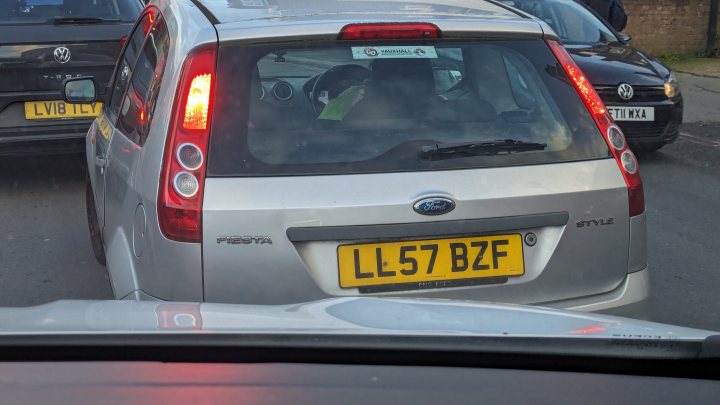 Pistonheads - The image features a silver hatchback car on a street, driving away from the camera. The car has a visible license plate that reads "L62 BZF". The vehicle is captured in motion, with its taillights illuminated. There are other cars on the road behind and ahead of it. The background suggests an urban setting, possibly during evening or nighttime, given the dim lighting conditions.