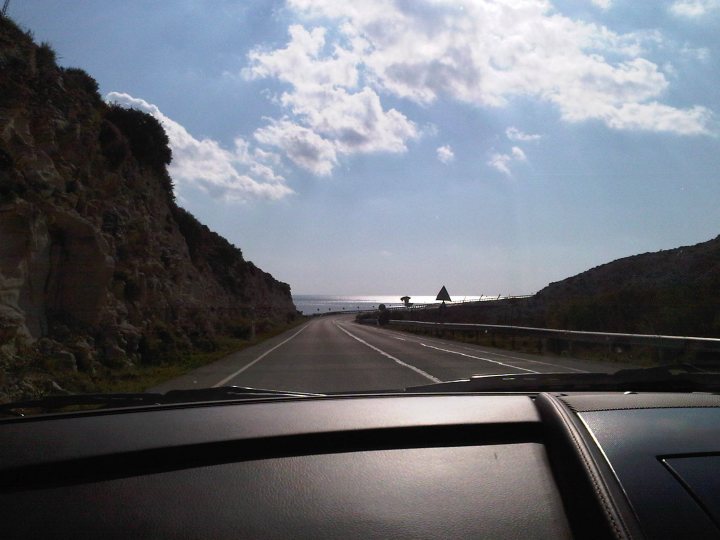 Hello from Cyprus! - Page 1 - Aston Martin - PistonHeads - The image captures a picturesque view of a highway that seems to stretch into the distance. The road is clear, bordered on both sides by rocky cliffs that offer a rugged charm. A body of water shimmers in the background, its surface reflecting the expansive sky above, where a few clouds might be on their way or have just passed by. This scene paints a serene picture of a day both on and off the road.