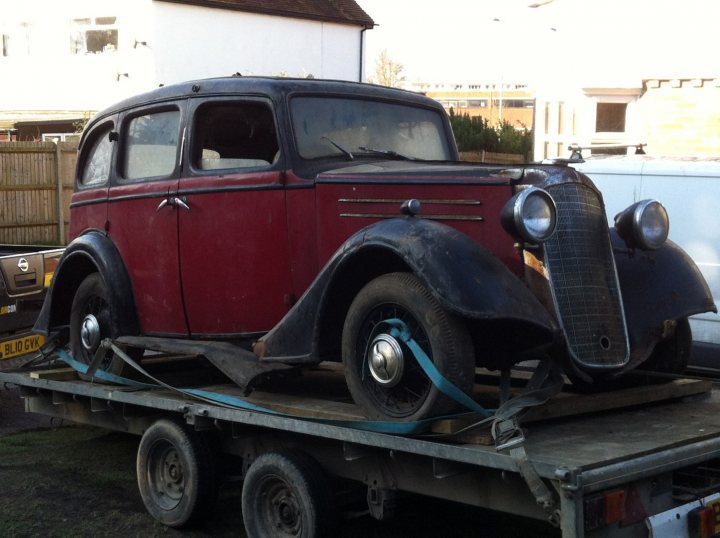 Show us your vauxhall! - Page 11 - VX - PistonHeads - The image shows an old, dark-colored car being hauled on a trailer. The car has a vintage design with round body and beige-colored stripes. It's secured on the trailer by straps, and the front part of the vehicle is visible. The trailer is positioned on a street, and there are other vehicles parked throughout the scene. The overall setting is urban, with a clear sky suggesting fair weather.