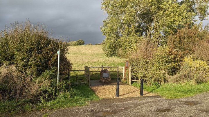 Pistonheads - The image depicts a rural scene, dominated by a stone building on the left side. In front of this building is a dirt road, which leads to a wooden gate in the center of the frame. Beyond the gate, there's a grassy field that stretches into the distance. On the right, there's a single tree standing alone, its lone presence contrasting with the open space around it. The sky overhead is overcast, suggesting a cool or gloomy day.