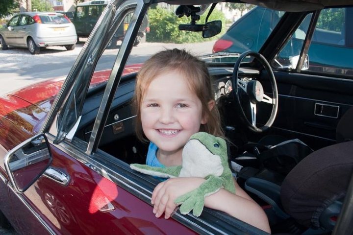 A little girl sitting in a car seat with a teddy bear - Pistonheads - In the image, a young girl is sitting inside a red car. She is holding a green stuffed frog. The car seems to be parked, with the steering wheel visible in the image. There are other cars in the background, indicating that the car is parked in a public setting like a parking lot. Her smile suggests that she is content and possibly excited about her car ride or her new toy.