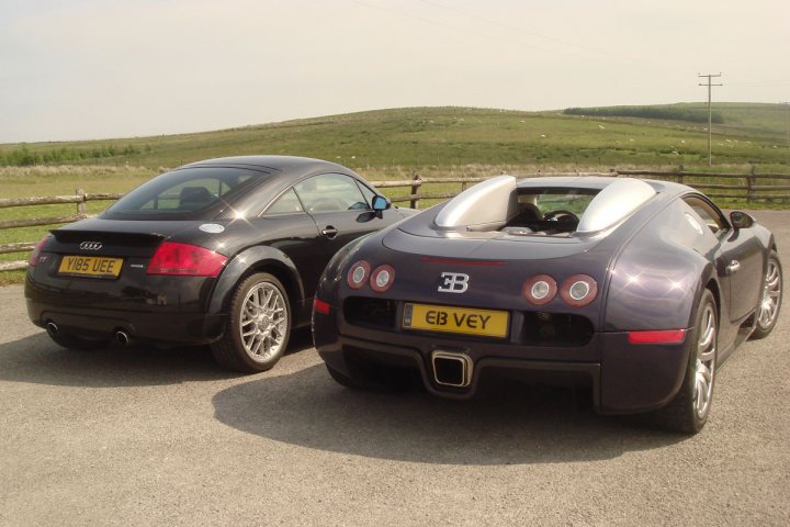 RE: Bugatti Veyron 16.4 | PH Used Buying Guide - Page 1 - General Gassing - PistonHeads UK - The image presents a tranquil scene of two sports cars parked side by side on a gravel surface. The car on the left is black, while the one on the right is blue. Both cars are facing towards the camera, showcasing their sleek designs and shiny surfaces. They are parked in front of a grassy area, adding to the serene atmosphere. In the background, there's a fence and trees, providing a natural backdrop for this automotive display. There's also a lone figure visible near the cars, perhaps indicating human activity or interest in the vehicles.