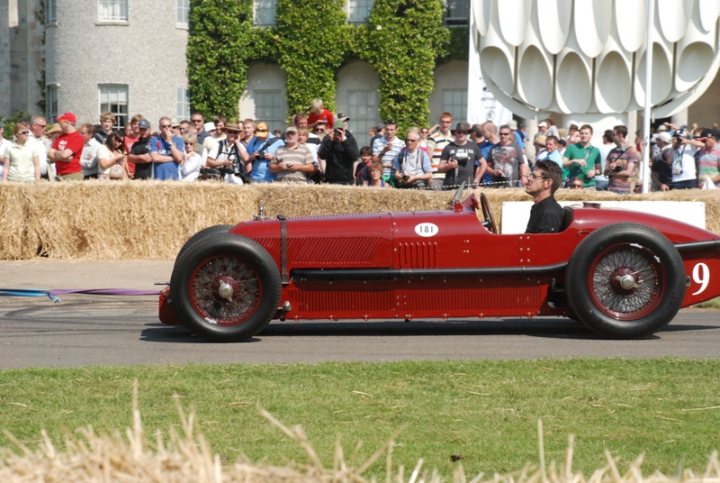 Please Help - Page 1 - Goodwood Events - PistonHeads - The image captures a vintage red race car on a track, possibly part of an event. A driver is seated in the car, wearing a helmet and goggles. The spectators, dressed in casual attire, line both sides of the track, watching the race car intently. Several have cameras, indicating that this is a special event. In the background, there are buildings with greenery, suggesting that the track is part of a larger sports complex.