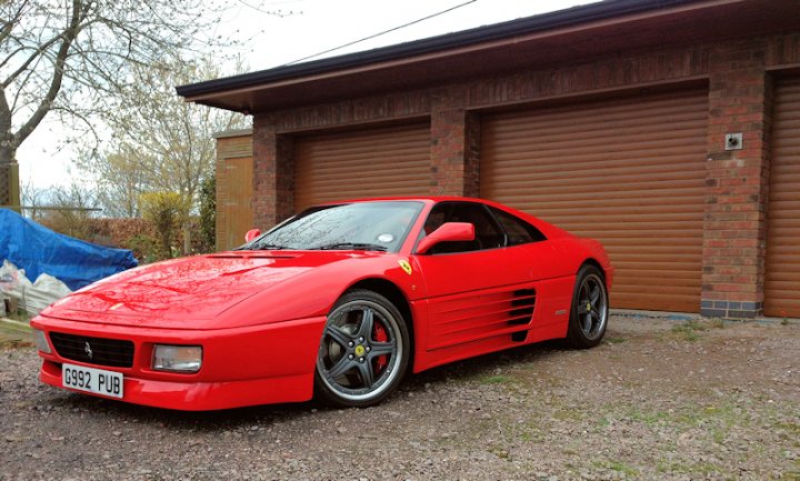 Ferrari 348 chit chat - Page 1 - Ferrari V8 - PistonHeads - The image features a striking red Ferrari parked in front of a garage with closed doors. The car is positioned at an angle to the camera, showcasing its sleek design and the Ferrari emblem on the front grille. There's a dusting on the ground, suggesting the car hasn't been driven recently, and a blue tarp can be seen behind the vehicle, possibly covering a work area or storage items. The overall scene conveys a sense of luxury, speed, and possibly a restoration or customization project.