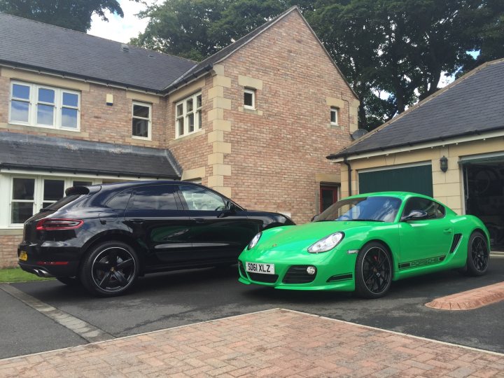 Macan GTS - Page 1 - Front Engined Porsches - PistonHeads - The image shows a residential driveway with two distinctive cars parked in front of a house. The house is a two-story structure with visible brickwork on the upper levels and a single-story, lean-to structure on the left, which could be a garage or extension of the house.

The darker car is a sleek, blue-black model, possibly a coupe or sedan, with a glossy finish and large alloy wheels. Its design suggests it is a modern, European car. 

The car with the green and black design is a sports car, likely a Porsche given its distinctive shape and branding. It has a sloping rear window and a forward-sloping hood, indicative of its aerodynamic design for high-speed performance. 

Both cars have UK-style license plates, suggesting the image was taken in the United Kingdom. The driveway itself is paved with interlocking bricks, and the area is well-maintained. The overall impression is of a well-kept, upscale residential setting.