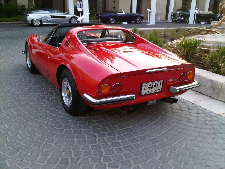 Dubai Pistonheads Mirage Royal Bonhams October Auction - The image features a sleek red convertible car parked on a gray gravel road that swirls around some paving stones. The car's top is down, and it has a license plate that reads "I 431". In the background, there are sculptures and other, less vibrant cars, suggesting that this might be a display area for luxury or classic vehicles. The overall setting conveys a sense of elegance and refinement.