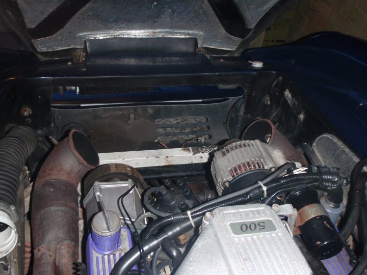 The image provides a close-up view of a car's engine compartment. The engine appears used, with signs of wear and maintenance. Numerous hoses and wires crisscross the space, connecting various components. The overall color scheme of the engine bay is a mix of metallic tones and the darker shades of the mechanical parts, suggesting a complex machine with multiple layers of functionality.