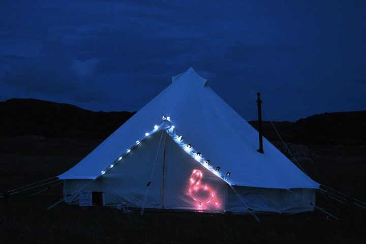 Anyone else got a bell tent ? - Page 1 - Tents, Caravans & Motorhomes - PistonHeads - The image depicts a large, white tent pitched in open field under a starry nighttime sky. The tent top has a distinctive wedge shape, reminiscent of a traditional teepee. Along the edge of the tent, a string of small white lights is stretched, lending an atmospheric glow to the setup. Marking the center of the tent is a luminous pink owl design, lending a touch of whimsy and uniqueness. The field seems to be in a remote area with small rocks and tufts of grass visible in the distance. The overall mood of the image is serene and inviting, suggesting an adventure or camping trip.