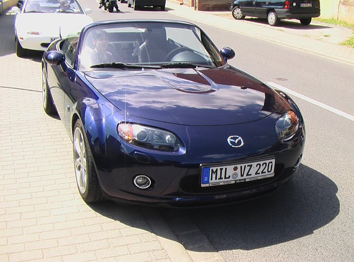 MK3 MX-5 for a 20 year old? - Page 1 - Mazda MX5/Eunos/Miata - PistonHeads - The image shows a blue Mazda MX-5 Miata sports car parked on the side of a street. The vehicle has a sleek design with a convertible roof and is positioned at an angle, allowing a clear view of its front. The setting appears to be an urban area as there are other cars and buildings in the background. The car's plate number "MI1 V20 56" is prominent, suggesting it may be a personal vehicle or used for test drives.