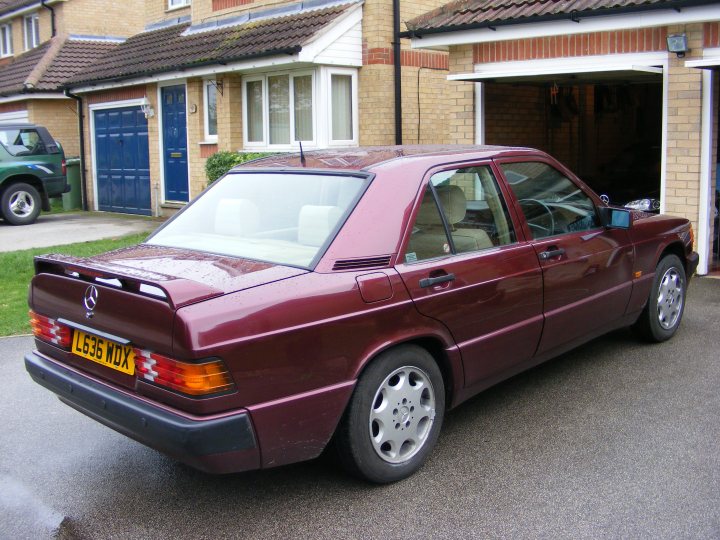 Pistonheads Short History - This image features a dark red or purple car parked in front of a house with a garage. The car has European plates and it is a manual transmission vehicle, as indicated by the gear column in the center console. The garage door is open, and another, newer car is visible inside. There is a driveway leading up to the garage, and the house has a brick facade with a blue front door, and a small green hedge. There are no visible people or animals in the image, and the street is not visible. The car appears to be in good condition, suggesting it might be well-maintained or recently purchased.