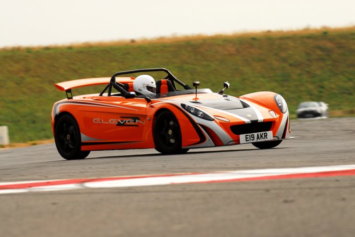 Road Legal 2-11 for the Track - Page 1 - Elise/Exige/Europa/340R - PistonHeads - The image depicts a single-seated orange and white sports car racing on a track. The driver is inside wearing a white helmet. The sports car is blurred as it moves, indicating speed. The background is out of focus, but some greenery can be seen, suggesting a rural or semi-rural track setting. The car number "19" and the words "GL- ????Owner" are visible on the side of the car.