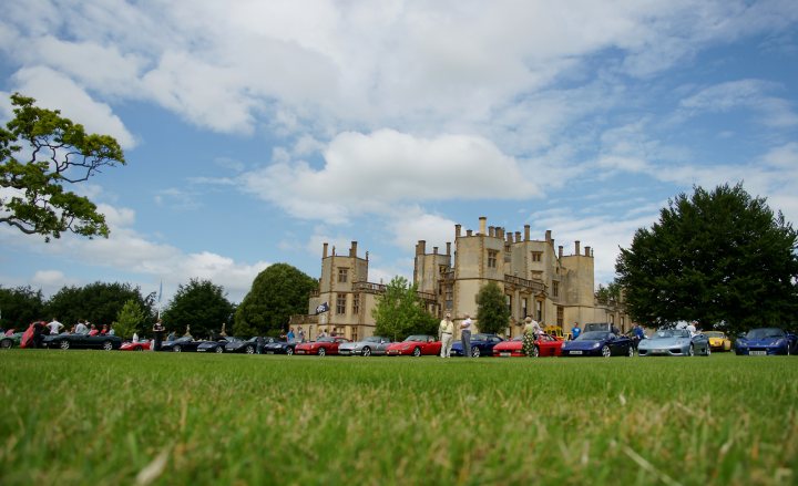 Pistonheads June Motors Rally July Manor Raleigh - The image captures a tranquil setting in an expansive field, where a line of vintage cars, spanning across several decades, are neatly arranged in a row, flanked by the backdrop of a classic castle-like building. The clouds in the sky provide a contrast to the clear blue of the day, adding depth to the expansive field. The green grass of the field gently undulates, creating a serene atmosphere.