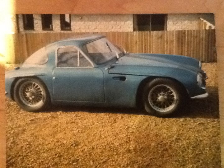 Early TVR Pictures - Page 10 - Classics - PistonHeads - The image is a vintage photograph of a small blue car captured from a non-front position, giving a side view. The car appears to be an older model, possibly from the mid-20th century, and has a classic style with a prominent grille and round headlights. It has two doors and is parked on a gravel lot with a wooden fence in the background, which suggests a domestic or rural setting. The photo has a faded appearance, typical of aged documents or photographs, which adds to the vintage feel of the scene. There are no visible texts or distinctive brands that can be identified confidently in the image.