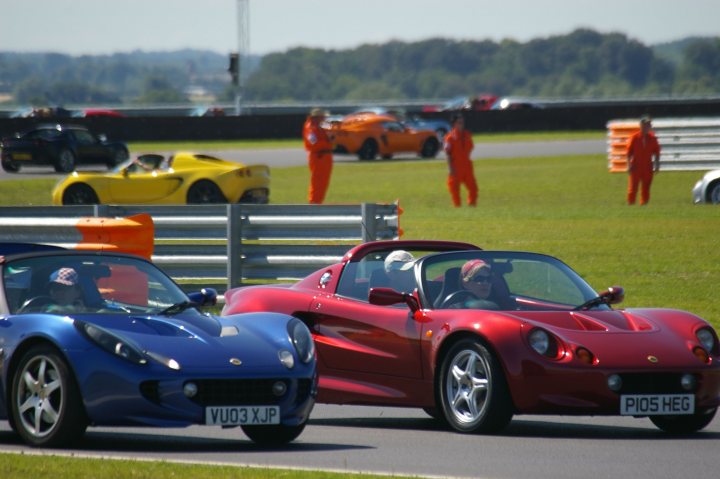 Snetterton Lotus Festival 25/26 June - Page 1 - General Lotus Stuff - PistonHeads - The image depicts a racing environment, with two sports cars situated on a track. The car on the left is blue, contrasting with the red car next to it. There are individuals wearing full orange racing suits standing in the background, likely with roles related to the event. The overall atmosphere suggests a high-performance event or competition.