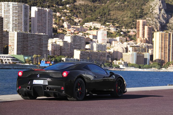 458 Speciale - Page 1 - Ferrari V8 - PistonHeads - The image features a sleek black sports car parked by the seaside, with the vibrant cityscape and a body of water in the background. The car has a white license plate and prominently displays the number 308. The cityscape includes tall buildings that appear to be residential, with lush green trees dotting the landscape. The sky is clear, reflecting off the surface of the water, suggesting a sunny day. The overall scene conveys a sense of luxury and tranquility.