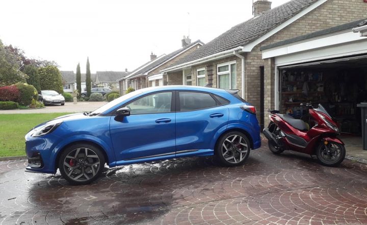 Pistonheads - The image shows a scene from outside a house. A blue sedan is parked next to the house, and it appears that it was recently rained, as there are water puddles around the car. There's also a red scooter parked in front of the car. A garage door can be seen in the background, partially open. The house itself has a chimney visible on its roof, suggesting that heating might be used during colder months.