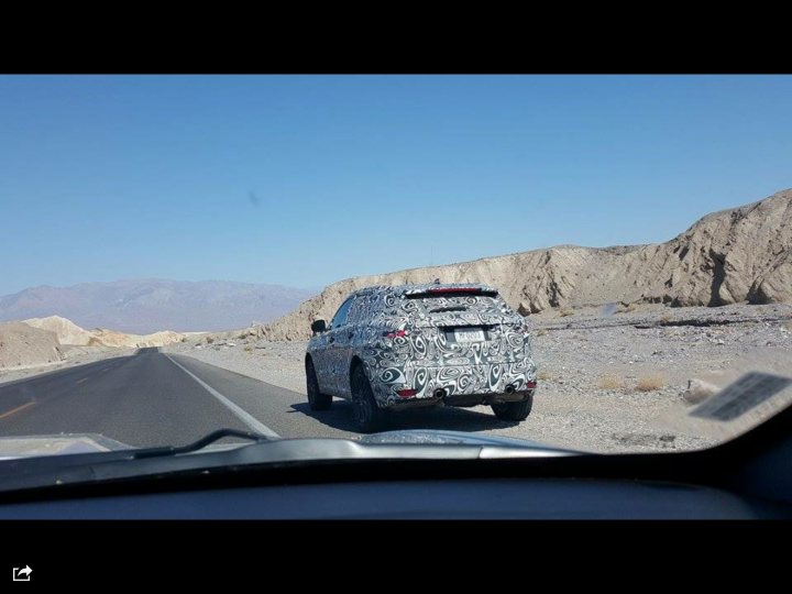 New Jag 4x4 spotted - Page 1 - Jaguar - PistonHeads - In the image, an SUV with a camouflaged wrap on its side cover is driving down a desert highway, passing a rocky outcrop to the right. The sky above is clear and blue, creating a vast open landscape that contrasts with the vehicle's camouflage design. There are mountains visible in the distant background, adding depth to the scene. The overall atmosphere suggests a journey or exploration in a remote, desert environment.