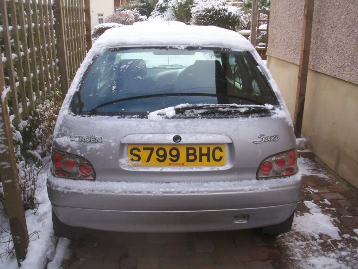 Saxo Pistonheads Citroen - The image captures a scene of a silver car parked in a driveway after a spell of snow. The car, slightly dusty, is covered with a layer of snow on its roof and hood, indicating that it's been parked there for a while. The license plate stands out against the snowy car with its bold yellow color and black text. A trail of the white salt used for anti-icing is visible on the ground, hinting at the recent activities in the area. The car is positioned against a backdrop of a calm and quiet residential setting.