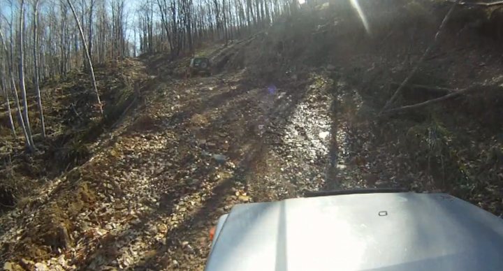 Bit of off-roading, and a bit of camping. - Page 1 - Off Road - PistonHeads - This image is a photograph taken from a forest trail. The trail is rugged, with uneven terrain and areas of loose soil. The sunlight is streaming through the canopy, illuminating the forest floor and creating interplaying shadows across the path. The vegetation surrounding the trail appears lush, with various trees and bushes. The bottom section of the image is partially obscured by a metal object, but it doesn't provide any significant context. There are no human figures or identifiable landmarks in the photo, suggesting it might have been taken for the purpose of showcasing a natural scene or during an outdoor activity like off-road driving.