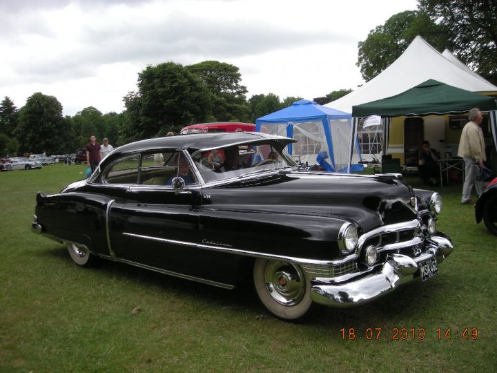 Classic Pistonheads Spotters - The image features a black classic car prominently displayed on a grass field. This vintage car, seemingly an antique, has a very shiny surface that gleams in the daylight. The setting is a grassy area where multiple classic cars and people can be seen. There are also a few umbrellas and tents, suggesting a possible outdoor auto show or gathering. The weather appears to be overcast, casting diffused light on the scene.