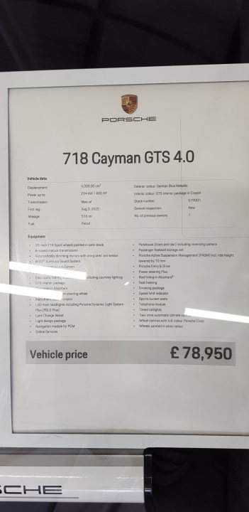 The image shows a framed document with text and pricing information. It is displayed on a wall or stand, with the frame's glass partially visible. The text and numbers suggest that this could be a vehicle sales listing, possibly from an auto show or dealership. There's also a partial view of a car model, as indicated by the "Porsche" logo in the background.