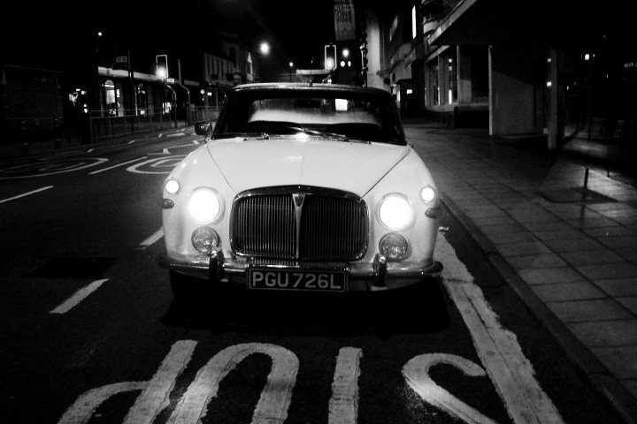 Evening and night photography - Page 4 - Photography & Video - PistonHeads UK - The image features a vintage car, specifically an early-model Rolls Royce, which is driving down a street at night. The car has distinctive chrome details, and the license plate reads "D79 MK." The setting appears to be urban, with the vehicle on what looks like a British road, as indicated by the road sign in the background. There's also a pedestrian crossing sign visible, suggesting that this is a regulated crossing area for pedestrians. The nighttime ambiance adds a sense of tranquility to the scene.