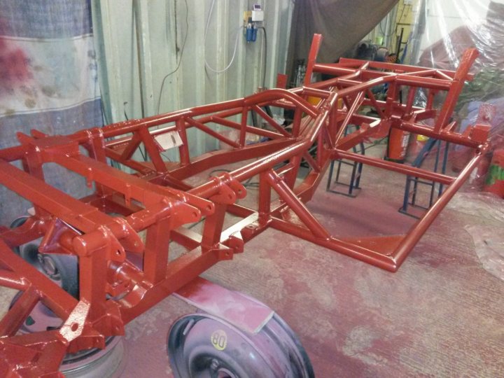 A body off in pictures, LOTS of pictures. - Page 1 - S Series - PistonHeads - The image showcases a bare residential basement that is being used as a workshop or garage. The main feature is the assembly of a red custom vehicle chassis which has been newly painted and is in the process of being put together. The primary structure consists of a vibrant red tubular frame with multiple support braces. The chassis seems to be a custom-made contraption rather than a typical finished vehicle. Surrounding the chassis, the workspace is littered with various tools, parts, and possibly other materials typically found in this type of environment. There is also a male figure wearing a white t-shirt towards the lower-right portion of the photograph, partially obscured by the objects around him.