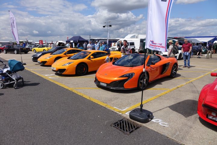 A bunch of cars are parked in a lot - Pistonheads - The image shows a lively car show taking place in a large parking lot. The main focus is on a line of brightly colored sports cars, including vibrant orange and striking red models. These cars are neatly parked side by side, drawing the eye with their sleek designs and vivid hues.

The lot is bustling with activity, as a crowd of people can be seen walking around, admiring the vehicles on display. Their casual attire suggests a leisurely event, and some individuals are carrying backpacks and handbags, indicating they might be visitors or attendees of the show.

Numerous large billboards are scattered throughout the scene, providing information or advertisements related to the event or the cars being showcased. The overall atmosphere is one of excitement and admiration, typical of such gatherings where enthusiasts come together to celebrate automotive excellence.