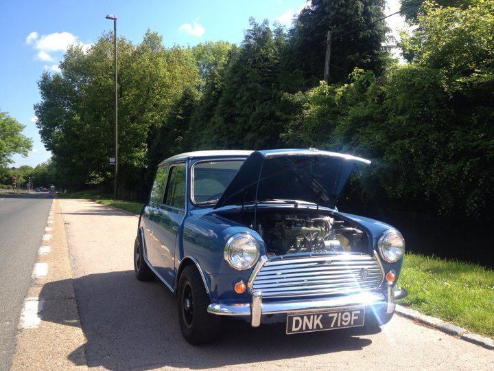 1968 cooper mk2. What wheels and tyres? - Page 1 - Classic Minis - PistonHeads - The image depicts a charming scene with a vintage blue Mini Cooper hatchback parked at the side of a road. The car has an open hood, revealing its engine to display engineering rather than to suggest a breakdown. The car is adorned with a license plate reading "DONK 719F". Behind it, the serene scenery includes lush trees framing an appealing countryside view. Above, the sky is clear, encouraging the occupants to enjoy a refreshing car ride on this sunny day.