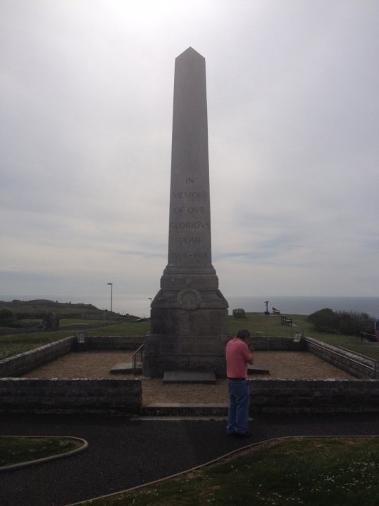 Where was I? Vol 3 - Page 25 - Holidays & Travel - PistonHeads - The image captures an individual standing at the base of a large, imposing stone monument on a desolate, empty field near a coastline. The monument is prominent, featuring an inscription at the top, and is set against a backdrop of a cloudy sky and a vast body of water. The person pauses, momentarily taking in the tranquil surroundings under a grazing tom cat. The scene exudes a sense of solitude and quiet reflection.