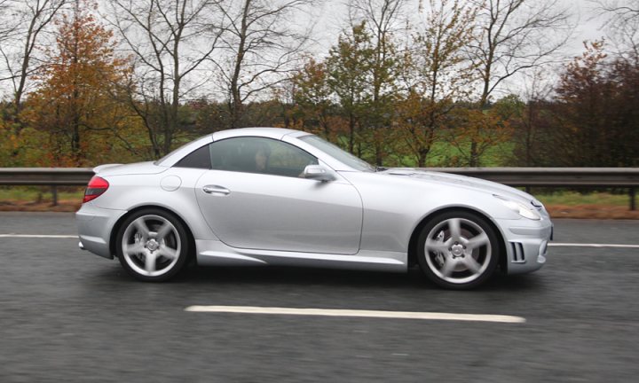 Pistonheads - The image shows a silver convertible sports car traveling down a road. The car has a sleek design with a soft top roof. The surrounding environment is a natural landscape featuring bare trees and what appears to be a grassy area. The road curves to the left, and the car is seen in the middle of the lane. The lighting on the car suggests daylight, and the weather seems to be overcast.