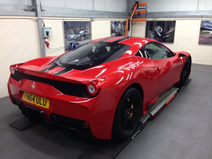 Our Cars - Page 135 - Scotland - PistonHeads - The image displays a red Ferrari sports car parked in an indoor garage. The car's sleek design and shiny paint suggest it's well-maintained. Its black wheels and rear spoiler enhance the vehicle's aerodynamic characteristics. The interior of the garage is partially visible, with a gray floor and some posters adorning the walls. Background details include metallic orange and black lifting equipment, indicating the car might be in a workshop or showroom. The top of the image shows a corner of a wall-mounted air vent, which suggests a clean and well-ventilated space.