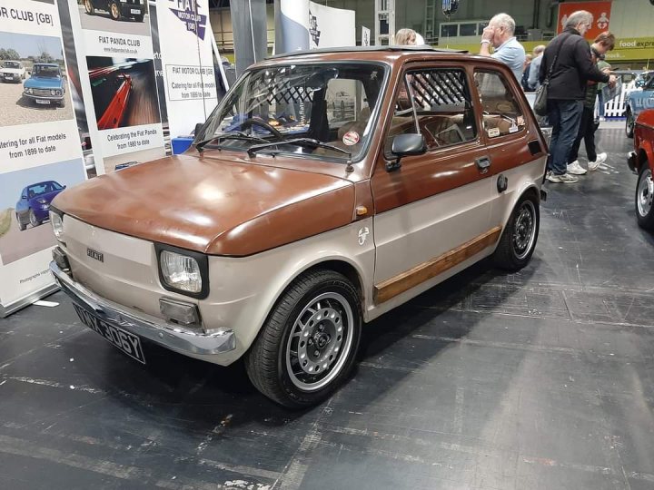 A car parked in a parking lot next to a car - Pistonheads - The image shows a classic car displayed indoors. It's an old, brown hatchback with distinctive silver rims and a noticeable rusted hood. The vehicle appears to be on display at a car show or exhibition, as indicated by the signage and other cars in the background. The overall setting suggests a casual event where enthusiasts gather to admire vintage and unique automobiles.