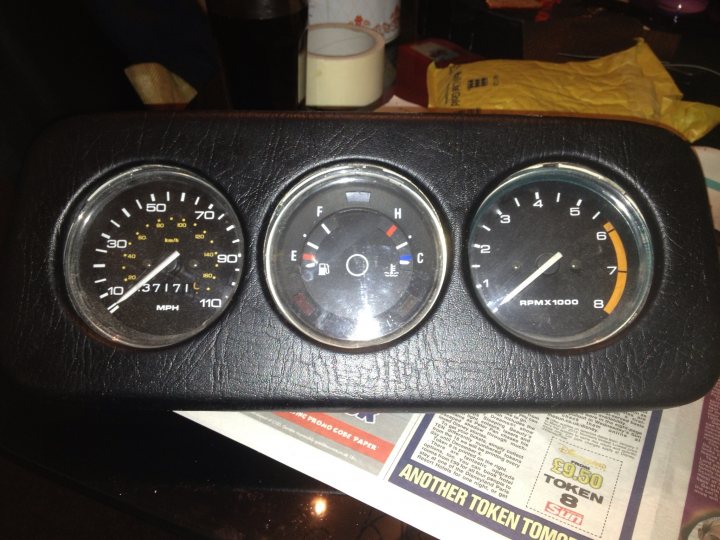 Pistonheads - The image showcases a unique three-panel car dashboard gauge resting on top of a newspaper. The gauge is predominantly black, with a cluster of white and yellow numbers representing RPM (Revolutions Per Minute). Two of the panels are black, while the center panel has a tachometer design. Additionally, there appears to be an auction notice for three tokens included in the newspaper under the gauge.