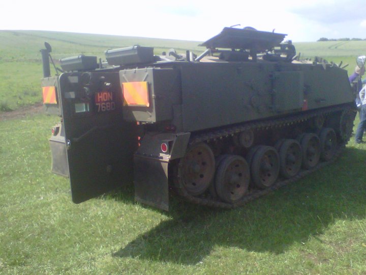 A good day out! - Page 1 - Off Road - PistonHeads - The image shows a large, green military tank parked on a grassy field. The tank is adorned with various features, including a cannon barrel, an emblem that reads "HON 7680," and a number of other markings and attachments. The tank is equipped with thick treads for movement and has a prominent angling at the loader's bay, as well as a series of wheels visible at the rear. The sky above is clear and bright, indicating fair weather conditions. There are two visible individuals in the background on the left side of the image, suggesting the presence of personnel or soldiers in the vicinity. The overall scene appears to be an open outdoor area, possibly a military base or training facility.