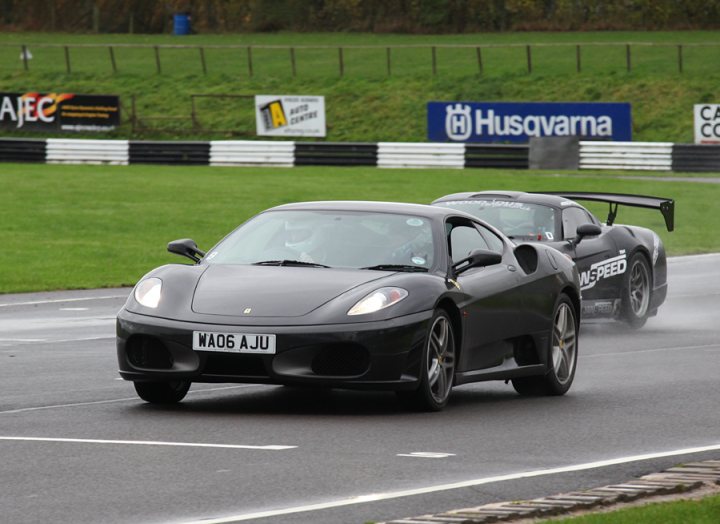 Pistonheads - The image shows a high-performance sports car on a racetrack, driving at high speeds as indicated by the water spray. Another similar car can be seen in the background, maintaining a safe distance. The car is sleek and has a distinctive emblem on its front. The surroundings include a grassy field and a fence.