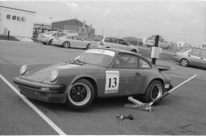 Anyone want some free photos of their cars? - Page 1 - East Anglia - PistonHeads - The image depicts an outdoor scene in what appears to be a parking area. A small, dark-colored sports car is the central figure, occupying much of the frame and positioned perpendicular to the direction of the lines on the pavement. The car is equipped with a racing number "13" on the side, and the name "Peter White" is visible, suggesting the car might be a race car or used for track days. A small sign next to the car reads "Benich Automotive," possibly indicating the business or location. In the background, there are various cars parked, although they are less distinct and are likely spectators or participants in a racing event, given the context. The monochromatic nature of the image suggests it might be an old photograph or styled to look as such.