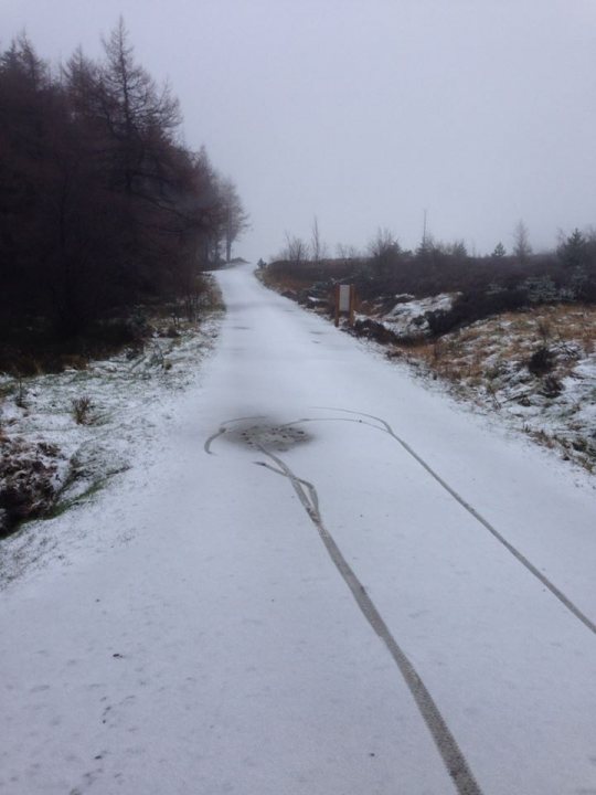 Pistonheads - The image depicts a wintery scene of a snowy road on a cloudy day. The road is lined with a rough, rugged terrain on both sides, suggesting a remote or rural area. A set of tire marks indicates recent vehicle passage, and a figure of some kind, possibly a pedal, is skidded on the road, indicating braking or a sudden stop. The sky is overcast, and the overall atmosphere is subdued due to the wetness of the road and the grayness of the sky. The harsh weather conditions are indicative of a cold, snowy day.