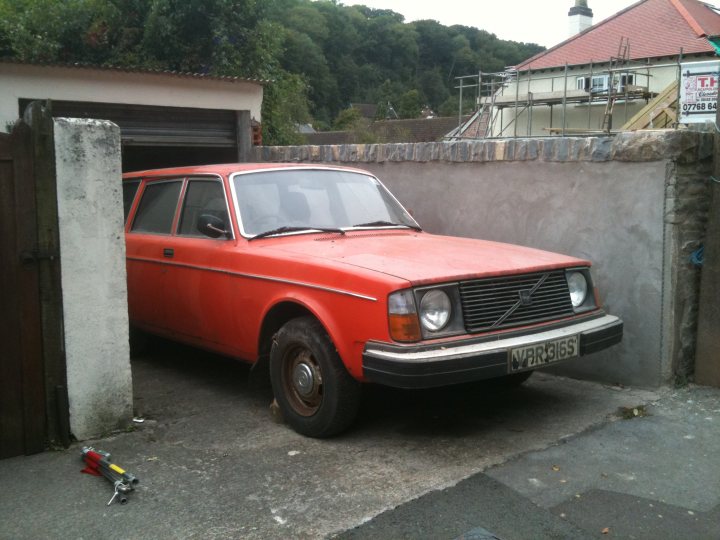 Pistonheads - The image shows an orange car parked next to a garage. The car appears to be an older model, possibly a Volvo or a similar brand, characterized by its distinctive design aesthetic. The vehicle features two doors, one of which is open, revealing a red interior. The garage itself has a white flat roof and is partially shaded by greenery. In the background, there is a partially constructed building with scaffolding, suggesting construction or renovation is taking place.