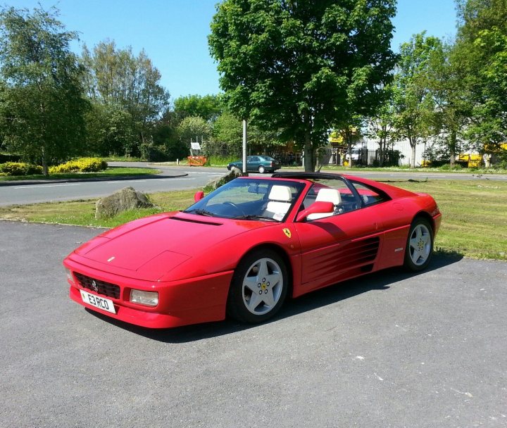 Ferrari 348 chit chat - Page 1 - Ferrari V8 - PistonHeads - The image shows a striking red Ferrari sports car parked on an asphalt surface. The Ferrari is positioned near a curb and is set against the backdrop of a grassy field and trees. The sky above is clear and blue, suggesting a bright day. There are some trees and a building in the blurred background, indicating an urban or suburban area. The vehicle appears to be well-maintained with clean, shiny bodywork and gold rims around the wheels.