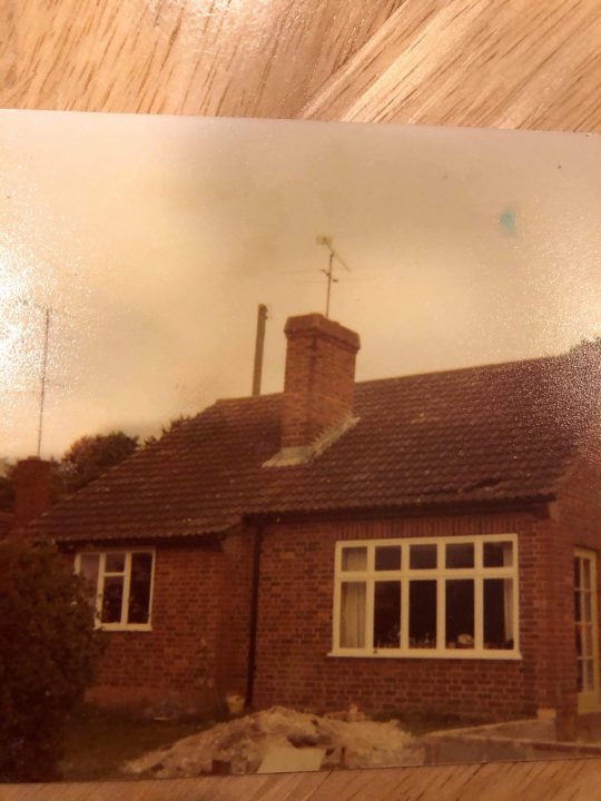 Build thread - bungalow to house - Page 9 - Homes, Gardens and DIY - PistonHeads UK - The image shows a physical photograph that has been slightly tilted. It depicts an old brick house with a visible chimney and a white exterior, possibly indicating age or maintenance needs. A small, flat-roofed extension is on the right side of the house. In front of the house, there's a driveway leading to what appears to be a single-car garage. The surroundings include a well-maintained lawn and some trees, suggesting a suburban setting. The photograph has a watermark or digital artifact across it that reads "2019," indicating when the photo was possibly taken or edited. The overall quality of the image is indicative of a personal snapshot rather than a professional real estate or architectural photograph.