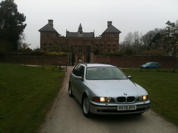 Pistonheads Photoshoot Locations - The image shows a silver BMW on the left of the road, approaching a wrought-iron gate that is part of a red brick wall. In the background, there's a large brick building featuring multiple spires and a central cross-like structure. The setting appears to be a countryside drive with grass on either side of the road, and trees lining the distance. The sky is overcast, suggesting it could be raining or just generally gloomy.