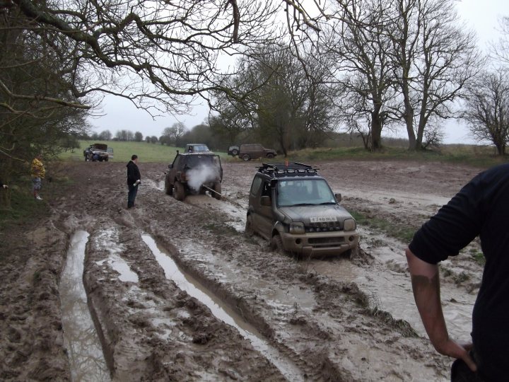 Pistonheads - The image captures an outdoor scene where a black vehicle is driving through a muddy field, with waterlogged terrain and scattered dirt. A person is standing on the left side of the field, observing or perhaps coordinating the activity. In the background, there are other vehicles and small trees indicating a rural setting. Despite the challenging conditions, the vehicles seem to be handling the muddy field effectively, albeit leaving significant tracks on the ground. The sky is cloudy, suggesting an overcast or possibly wet weather condition.