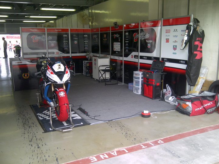 A group of motorcycles parked in a garage - Pistonheads - The image showcases a motorcycle racing setup. In the foreground, a red and white motorcycle is positioned, featuring a prominent number one and the logo of Repsol. A rider's gear is hanging to the right, indicating readiness for a race. In the background, a large, red and white partition with a promotional motorcycle display marks the beginning of the stall. The flooring is a striking red and white checkerboard pattern, adding to the overall theme. The setting suggests this might be a temporary garage or staging area at a motorcycle event.