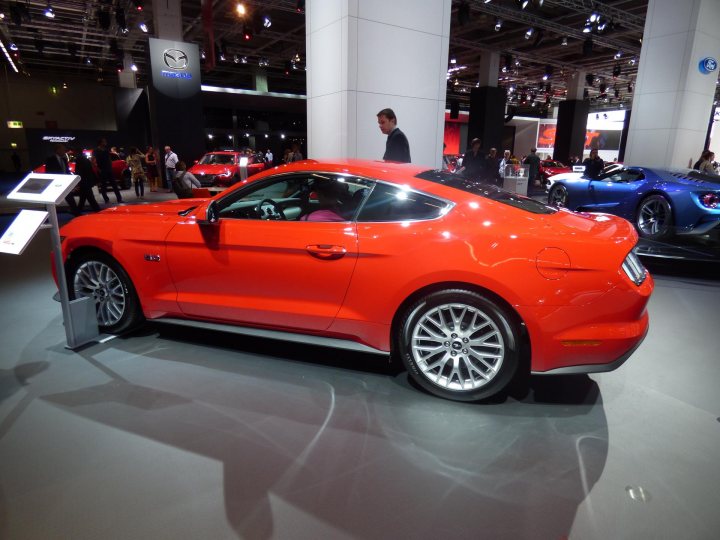 Mustang at IAA Frankfurt motorshow (pic heavy) - Page 1 - Mustangs - PistonHeads - The image captures the interior of a car show, where a vibrant red Mustang car glams against the center stage of a display. The Mustang is sleek and shiny, positioned on an angle to showcase its luxurious details. The floor beneath it is gray, providing a neutral background that contrasts with the car's vivid color. In the background, various people can be seen, some walking, others standing, all seemingly absorbed in the sights of the show. The overall setting is bright and well-lit, enhancing the appeal of the car and the environment.